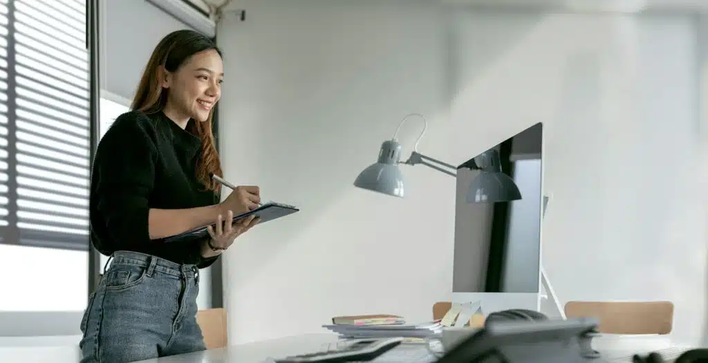 Smiley young businesswoman standing at desk, writing note and looking at monitor screen.