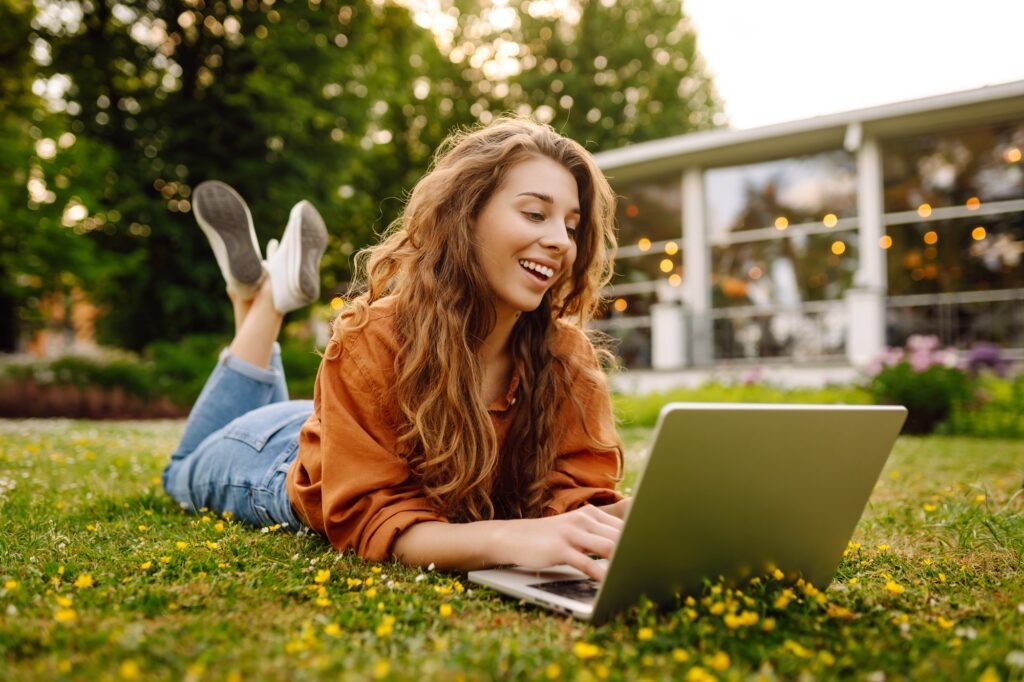 Beautiful female freelancer sits on green meadow with laptop. Online education. Freelancing concept.