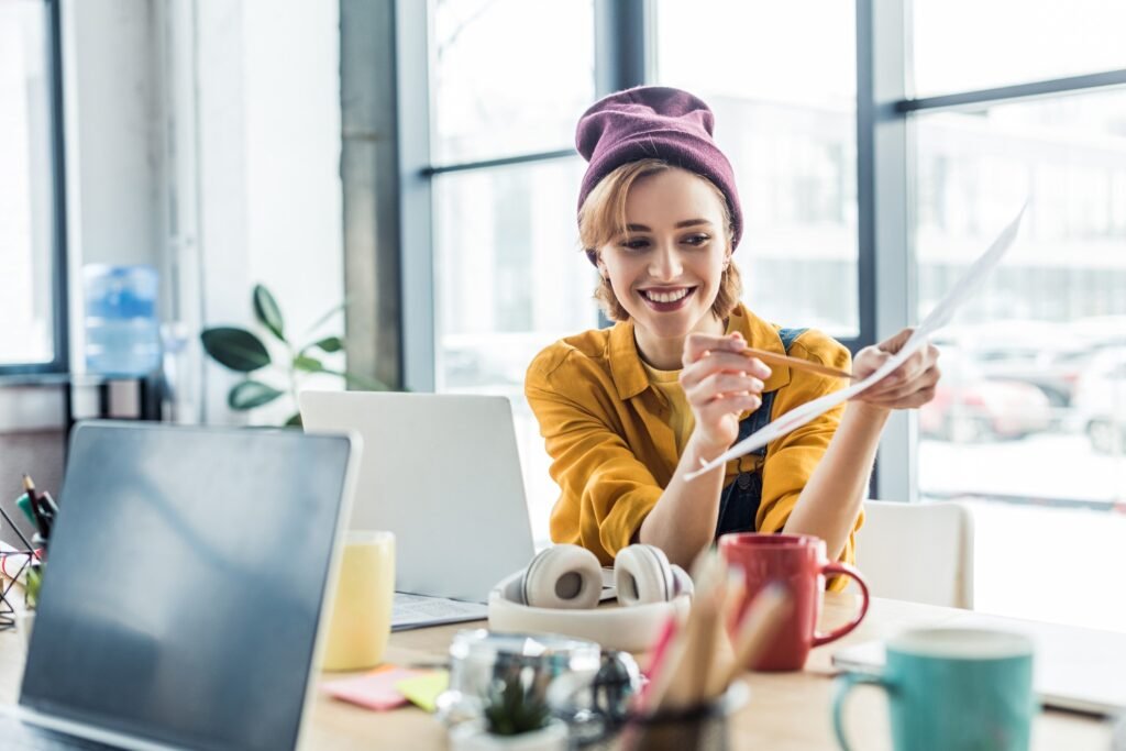 smiling young female it specialist holding document and pen while sitting at computer desk in loft