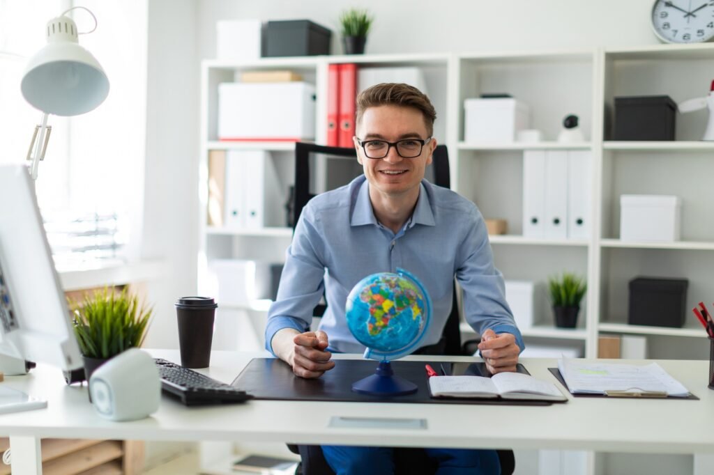 A young man sits in the office at a computer desk and in front of him stands a globe.