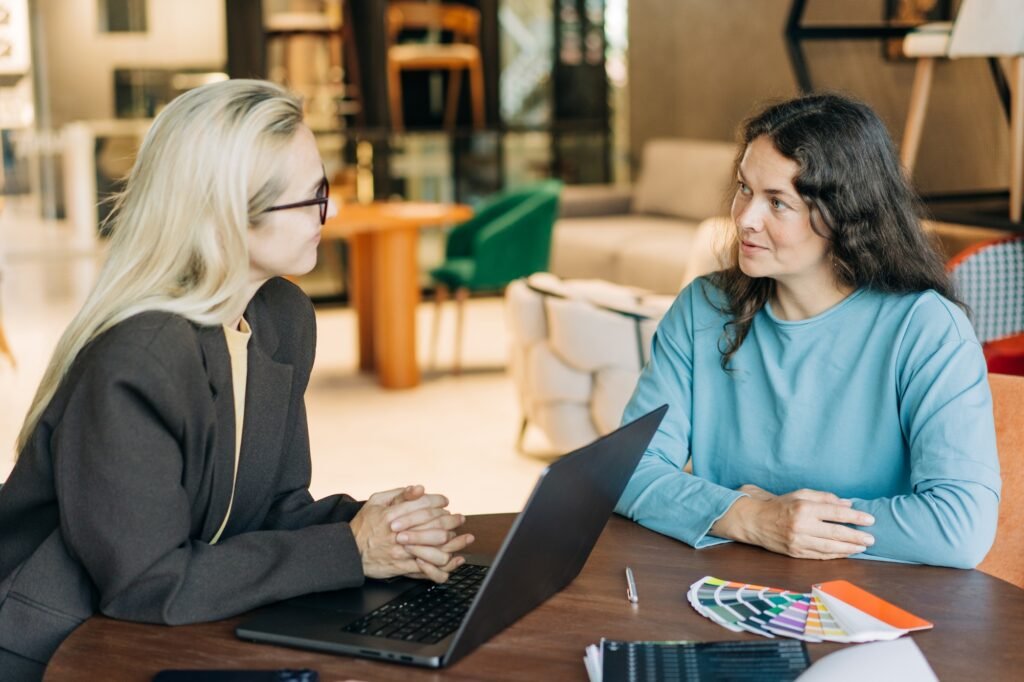 Two business women are talking while sitting at an office table in a furniture showroom.