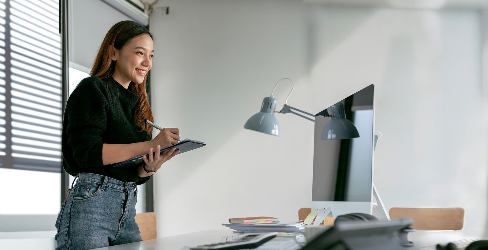Smiley young businesswoman standing at desk, writing note and looking at monitor screen.